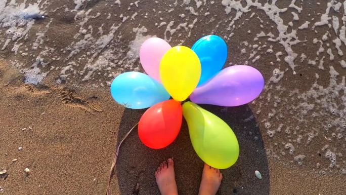 一束五颜六色的气球躺在海边的湿沙滩上，波浪翻滚，靠近一个人的脚。缓慢的运动。休闲娱乐娱乐休息娱乐庆祝