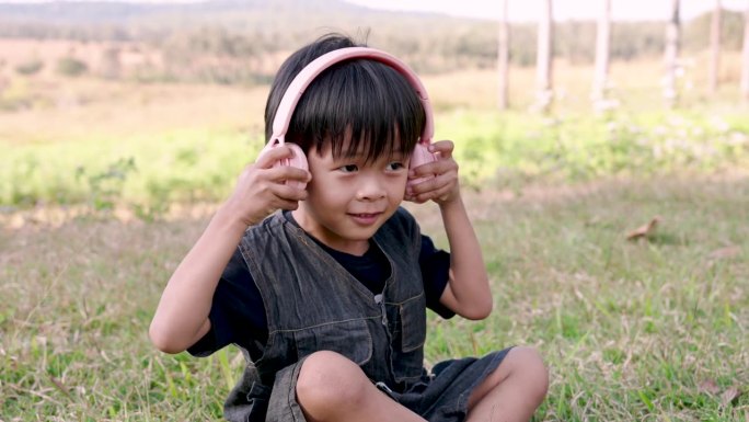 穿着深色工装裤的亚洲男孩盘腿坐在草地上，把耳机贴在耳朵上，脸上带着喜悦的表情。在阳光明媚的场地欣赏音