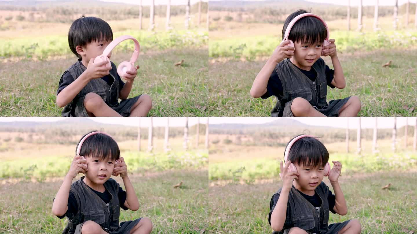 穿着深色工装裤的亚洲男孩盘腿坐在草地上，把耳机贴在耳朵上，脸上带着喜悦的表情。在阳光明媚的场地欣赏音
