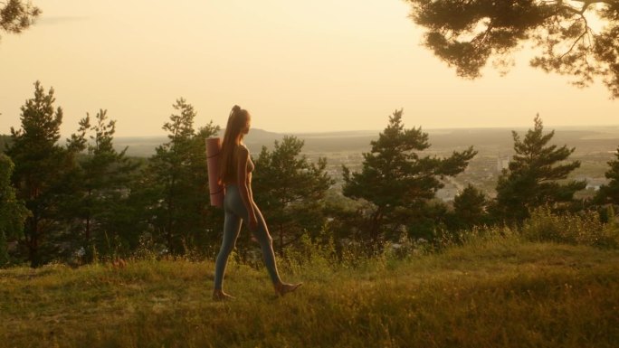 快乐的年轻女子穿着健身服，拿着瑜伽垫，在日落时分在森林里锻炼。