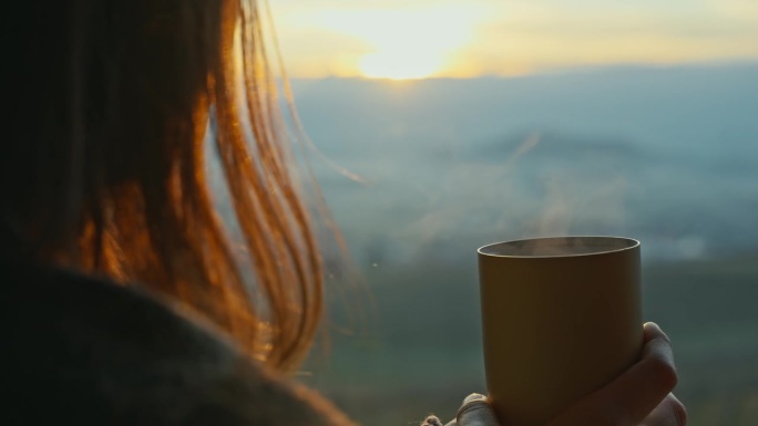 年轻女子手捧一杯热咖啡，享受窗外日落美景的特写后视图