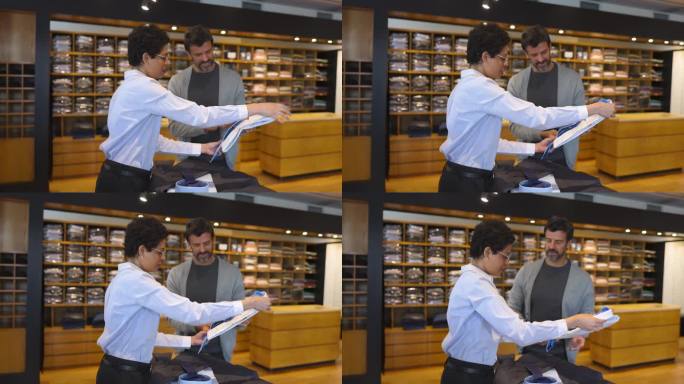 在一家男士商务服装店，混血女售货员正在帮助一位白人顾客挑选一件钮扣衬衫和领带