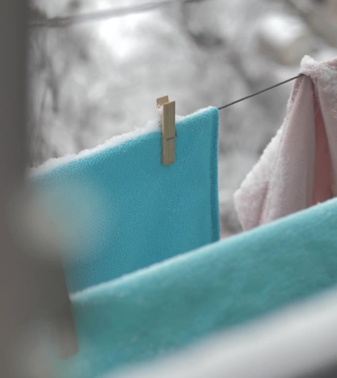 垂直:在下雪的寒冷的冬天，洗好的衣服挂在晾衣绳上。在寒冷的气候里晾干衣服。松软的雪落在奶油色、蓝色和