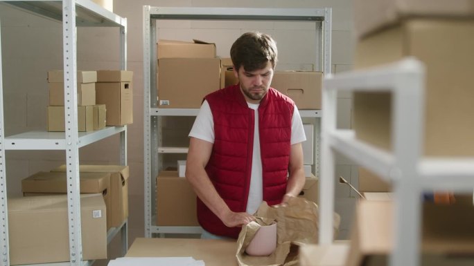 身穿红色马甲的男子，仓库员工将包裹装入投递箱，在纸板箱的背景下关闭并放在货架上。物流、配送和仓储概念