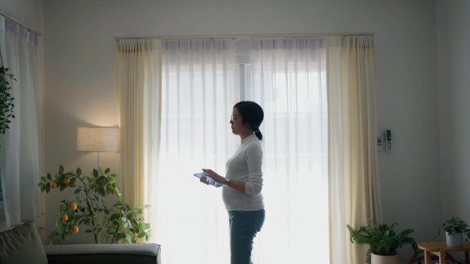 一位年轻的孕妇在家里的客厅里拿着平板电脑边走边说话。