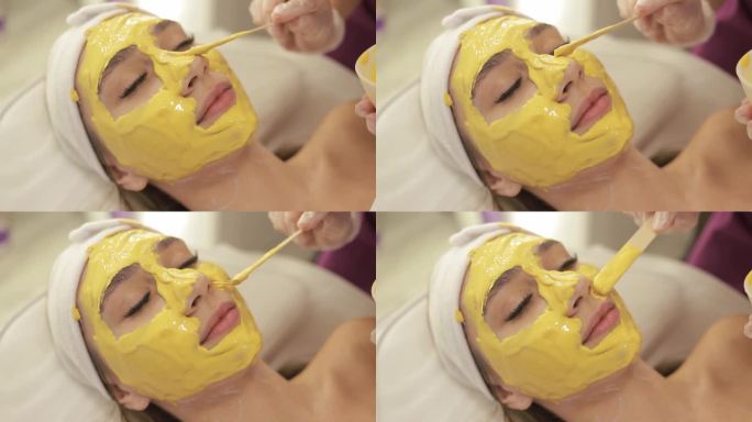 美容师用抹刀在一位女士的脸上涂抹金色海藻酸盐面膜。