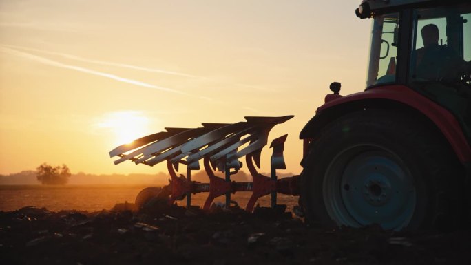 SLO MO男性农民在日落时分驾驶拖拉机对着天空犁地