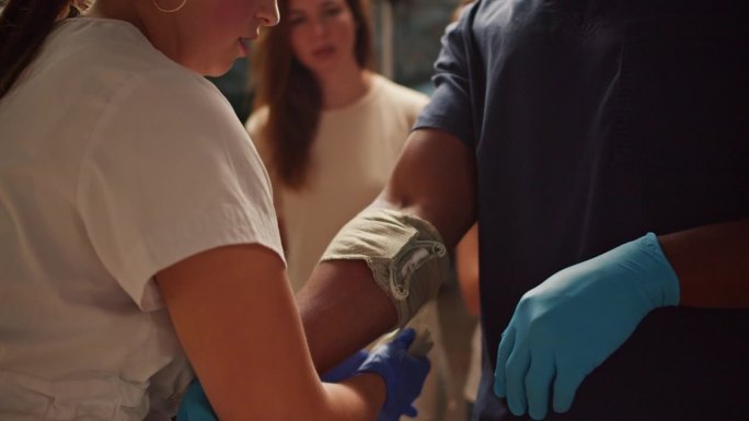 特写镜头:一名身穿白色医疗服的职业女护士在她的助手——一名身穿蓝色医疗服的黑人男子的手臂上缠上绷带。