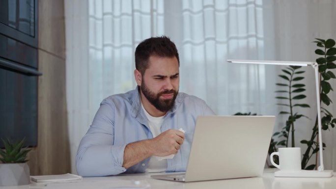 冷酷的黑发男人，流着鼻涕对着纸巾擤鼻涕，在公寓背景的电脑前远程工作。敬业的员工在病假期间在家回复商务