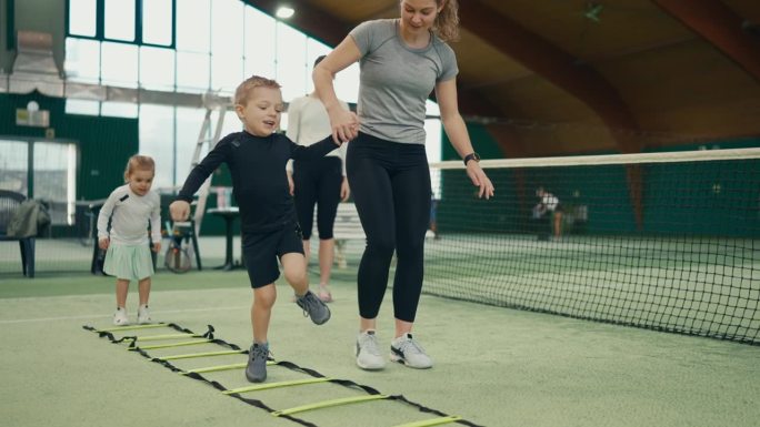 体育俱乐部网球场上女教练协助男孩进行敏捷阶梯训练