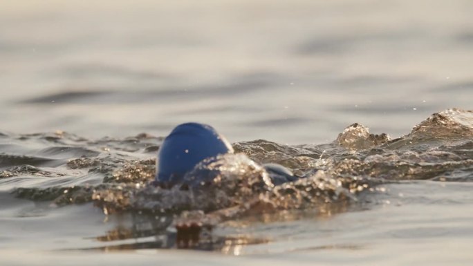 日落时分，戴着帽子在海洋中游泳的活跃女性的慢镜头镜头。一个女人正在海里游泳。