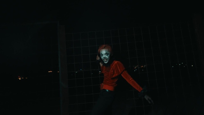 一名身穿红色衬衫、化着小丑妆的男子在铁栅栏前跳舞，在夜晚摆出令人毛骨悚然的姿势。