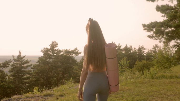 快乐的年轻女子穿着健身服，拿着瑜伽垫，在日落时分在森林里锻炼。