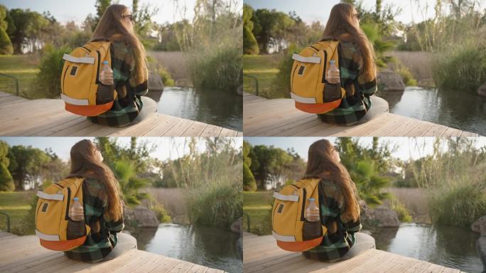 一个背着背包的孤独女人坐在一座木桥上，直视前方，深吸一口气，吸入新鲜空气，看着太阳。