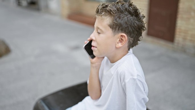 可爱的金发男孩自信地坐在城市的长椅上，一边观察户外，一边愉快地用智能手机聊天