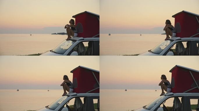 日落时分，女人坐在海边露营车顶上的帐篷旁