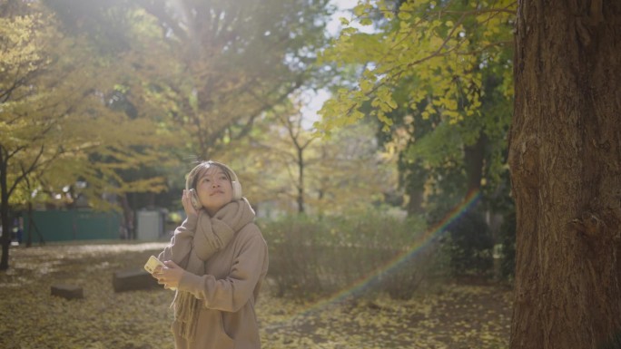 一个寒冷的秋天，年轻的亚洲女子在公园里放松。她用智能手机戴着耳机听音乐。