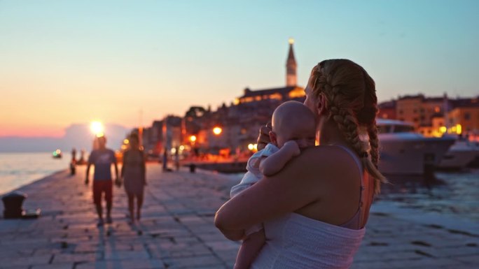黄昏时分，母亲抱着蹒跚学步的孩子站在城市的人行道上看风景。罗维尼，伊斯特拉，克罗地亚。