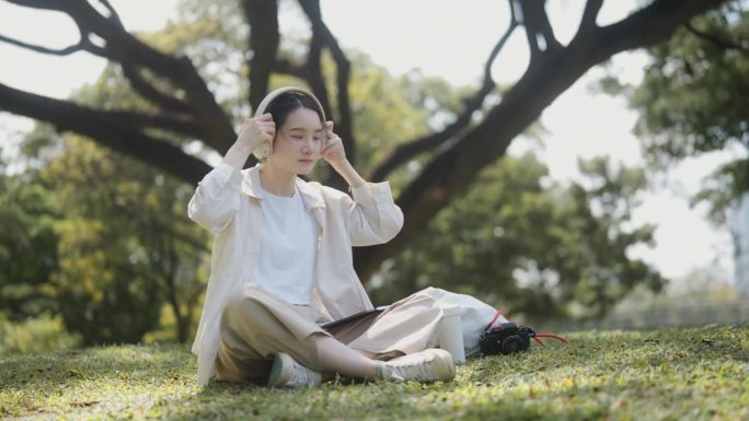 周末，亚洲青少年戴着耳机坐在草地上，玩着平板电脑放松。