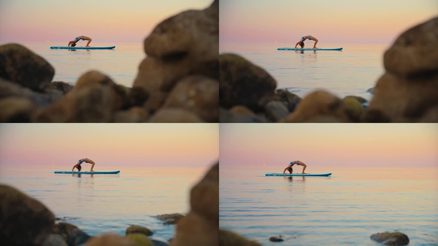 在日落时，健美女子在海上对着天空的冲浪板上练习瑜伽的慢镜头
