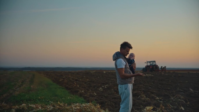 SLO MO Man农学家抱着小男孩，在日落时分用拖拉机对着天空犁地检查土壤。农田里的农民。