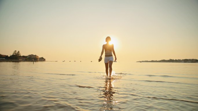 女子在度假期间在日落海滩浅水中散步的慢镜头
