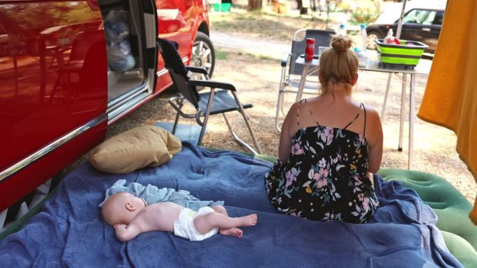度假时，母亲坐在露营车外床垫上熟睡的孩子旁边