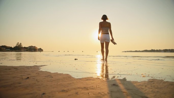 女子手持拖鞋在沙滩上漫步，欣赏日落的慢镜头镜头