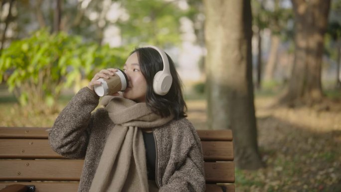 年轻的亚洲女子坐在美丽的风景公园在阳光明媚的日子。她正在用手机耳机听音乐，喝咖啡。