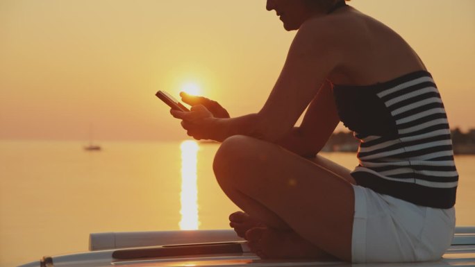 日落时分，一名女子坐在海边的露营车上使用智能手机