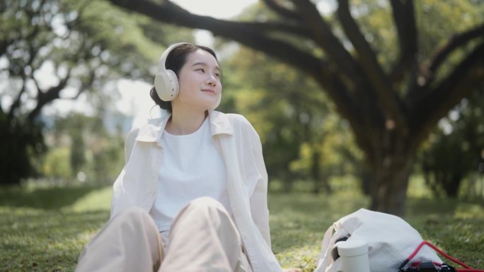 美丽的女人在阳光明媚的日子里坐在自然公园里，戴着耳机听音乐