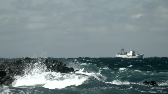 一艘拖网渔船在一个多岩石的海岸附近暴风雨的冬天航行