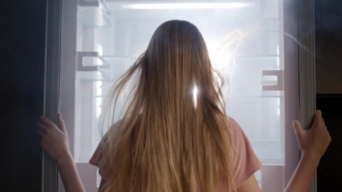 一位年轻女子向打开的冰箱里看，冰箱里有光，有雾。极度冷却和凉爽