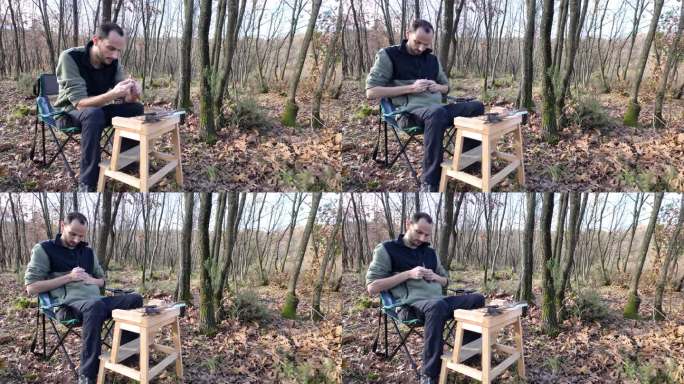 男露营者一边雕刻木雕，一边坐在满是秋叶的森林里的露营椅上。