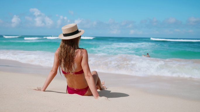 金发女子穿着比基尼草帽坐在沙滩