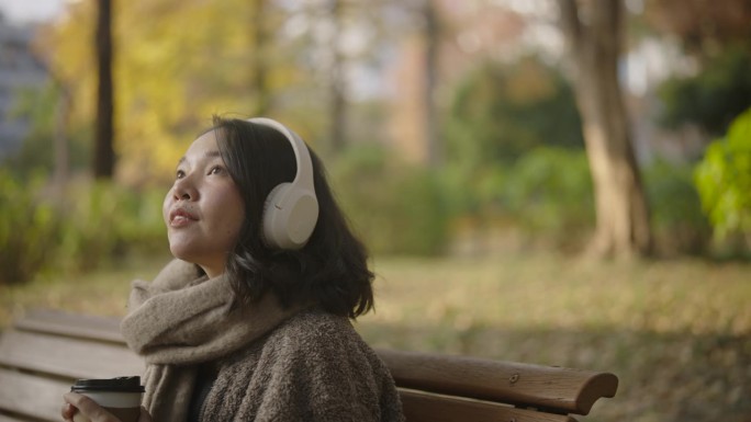阳光明媚的日子里，亚洲女游客坐在美丽的公园里。她边喝咖啡边用耳机听音乐。