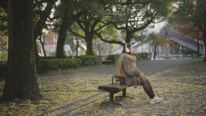 年轻的亚洲女子坐在美丽的风景公园在阳光明媚的日子。她正在用手机耳机听音乐，喝咖啡。