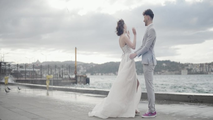 浪漫的新婚夫妇互相拥抱，站在码头上，面对着模糊的城市景观和绿色的山丘。行动。汹涌的海浪和新娘新郎的大