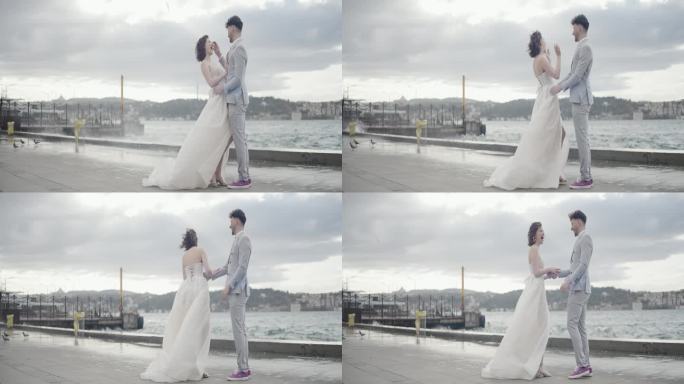 浪漫的新婚夫妇互相拥抱，站在码头上，面对着模糊的城市景观和绿色的山丘。行动。汹涌的海浪和新娘新郎的大