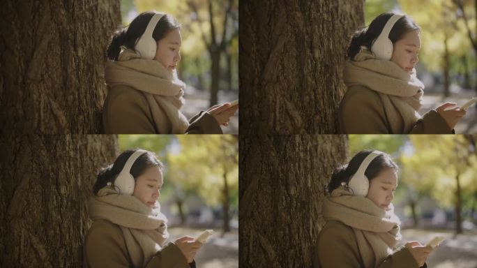 秋叶在日本。年轻的亚洲女子站在树下。她用智能手机戴着耳机听音乐。，享受放松。