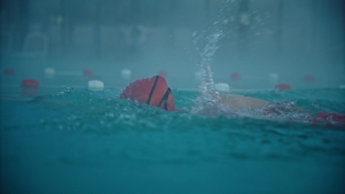 坚定的运动女子戴着帽子和护目镜在度假村的雾蒙蒙的游泳池游泳的SLO MO跟踪镜头