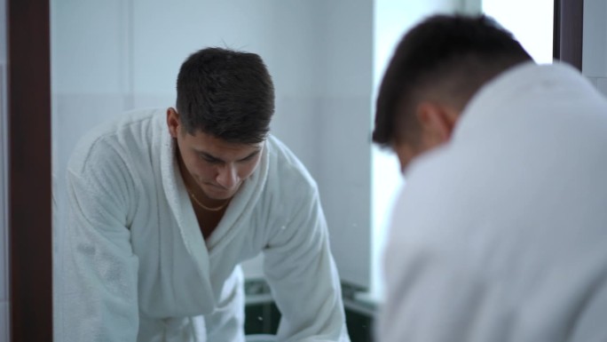 缓慢的运动。一个穿着白色浴袍的年轻人在浴室墙上的镜子前仔细地审视着自己。一个男人站在浴室里用清水洗脸