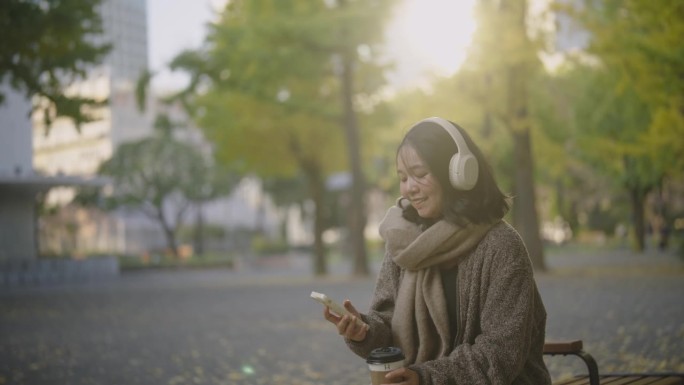 年轻的亚洲女子坐在美丽的风景公园在阳光明媚的日子。她正在用手机耳机听音乐，喝咖啡。