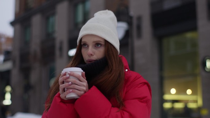 在寒冷的冬日里，戴着白帽子、穿着红色冬季夹克的冰冷的年轻女子在户外拿着外带杯里的热咖啡，喝着热咖啡。