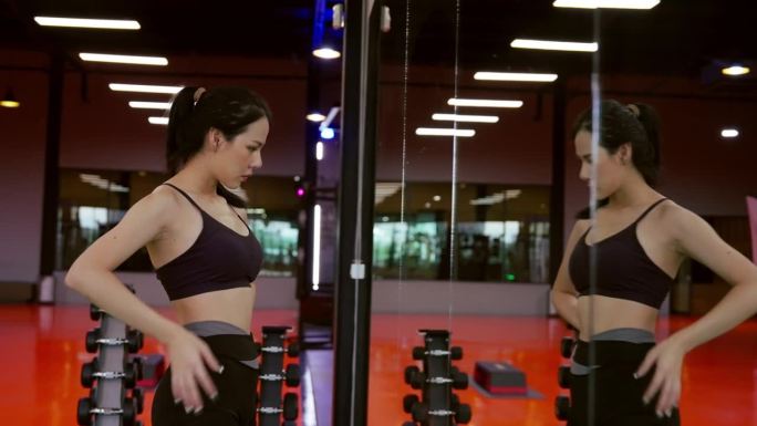 精力充沛的健身女运动员在健身房。亚洲女运动员站在窗前，看着我的身体。健身中心的美女美女和自信