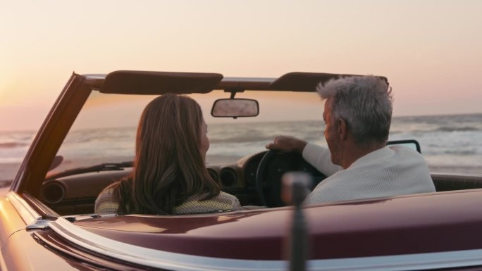 退休的高级夫妇度假坐在经典的敞篷跑车在海滩上观看早晨的日出公路旅行的慢动作拍摄