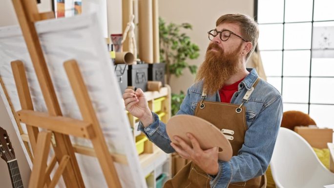 自信的年轻红发男子在艺术工作室里摇摆，微笑着让他的创造力发挥得淋漓尽致，手里拿着画笔，画着杰作