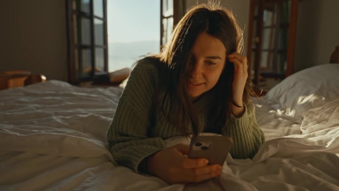 快乐的年轻女子从窗台上爬起来，倒在床上，在家里的卧室里使用智能手机