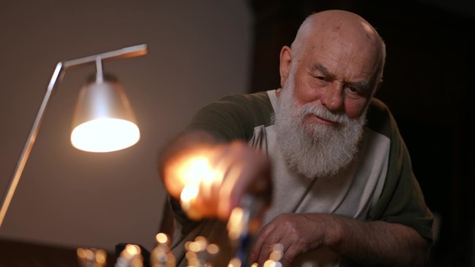 特写镜头。一个留着胡子的老人在一间黑暗的房间里，在一盏台灯的灯光下下棋。