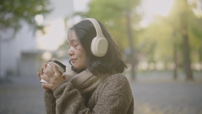阳光明媚的日子里，亚洲女游客坐在美丽的公园里。她边喝咖啡边用耳机听音乐。